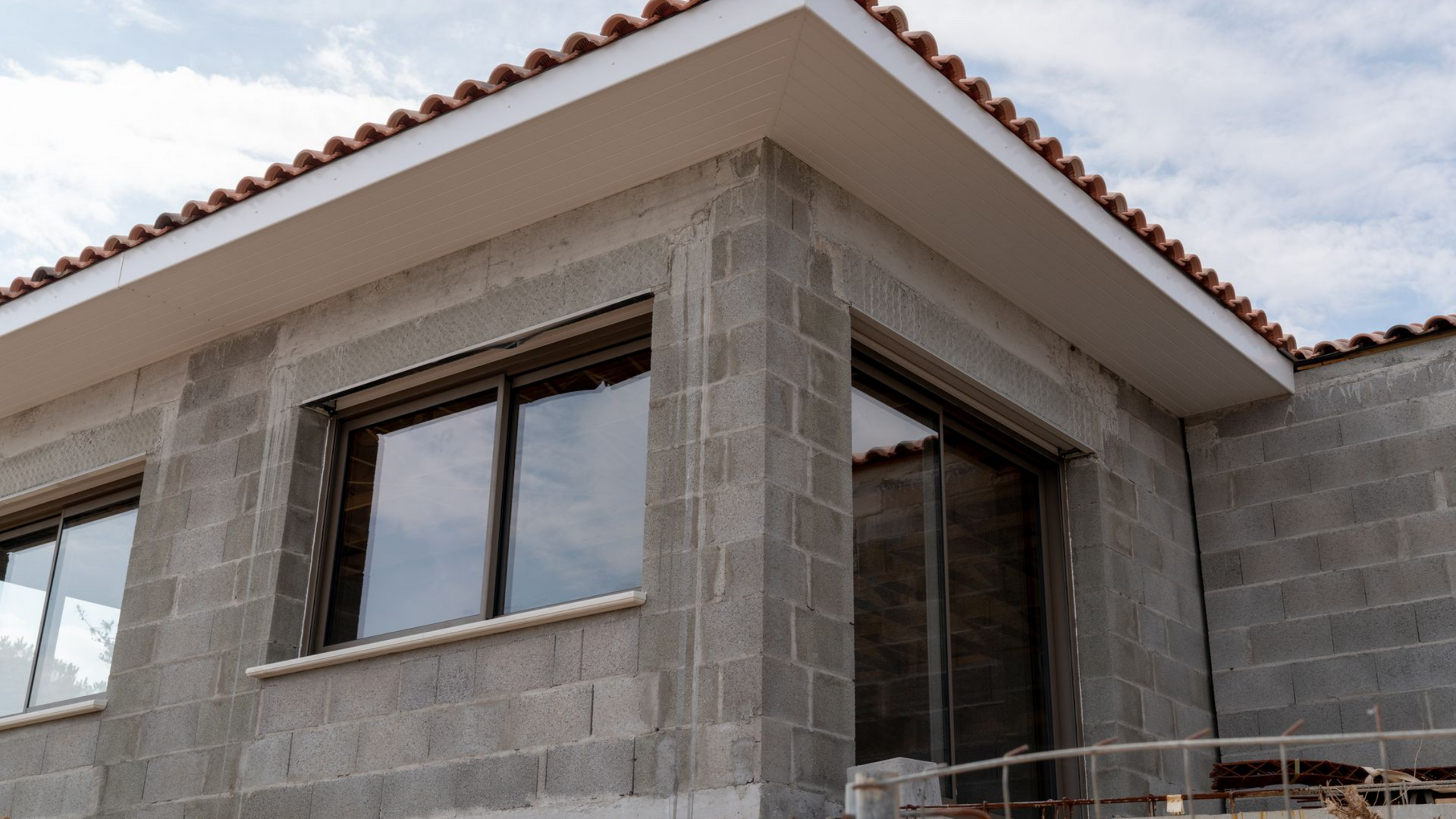 Angle extérieur d'un bâtiment en construction, avec des murs en parpaings gris, des fenêtres et un auvent blanc.
