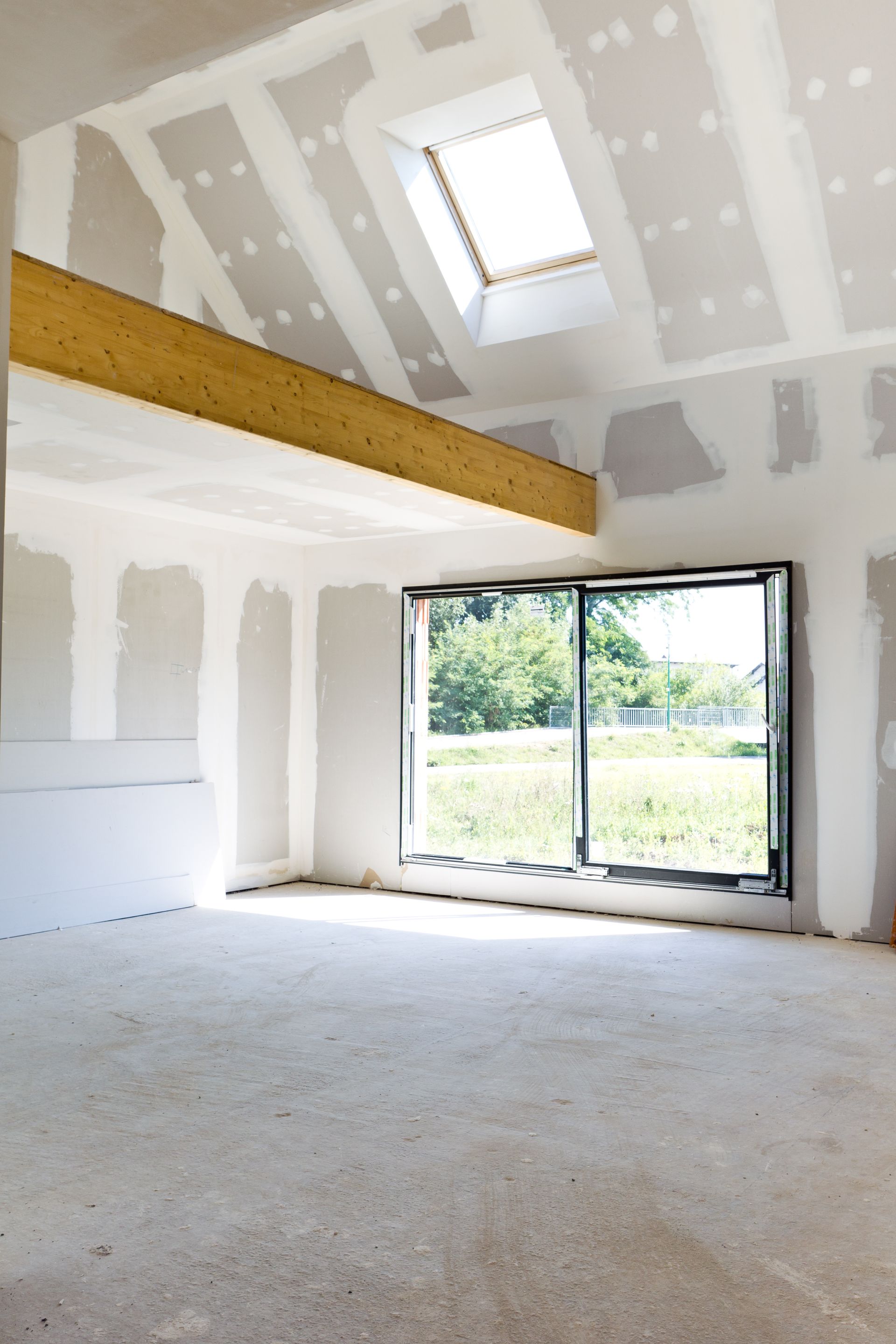 Intérieur d'une pièce en construction, avec cloisons sèches apparentes, puits de lumière et grande fenêtre donnant sur la verdure.