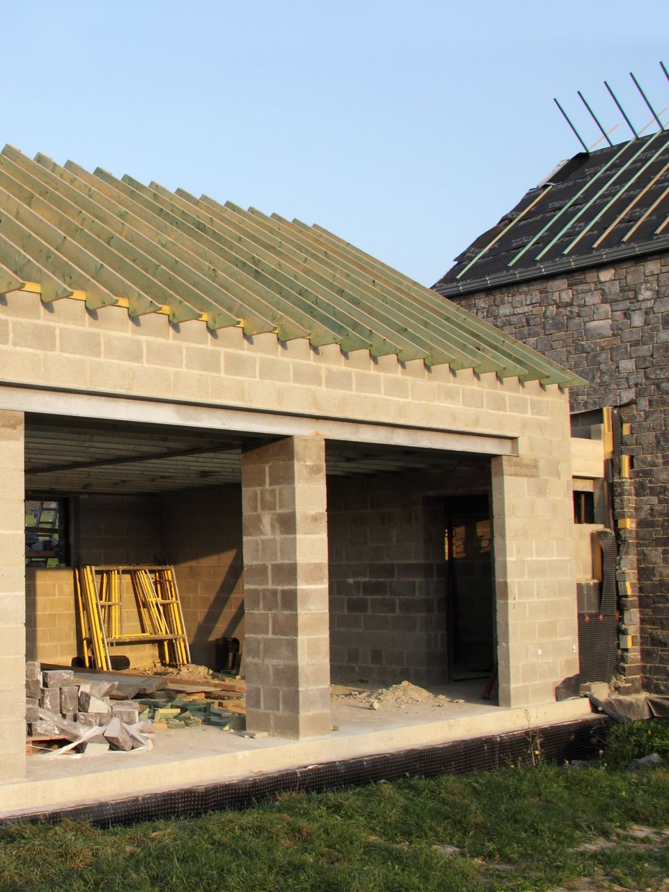 Bâtiment en construction : murs en parpaings de béton, toiture inachevée, structure en pierre attenante.
