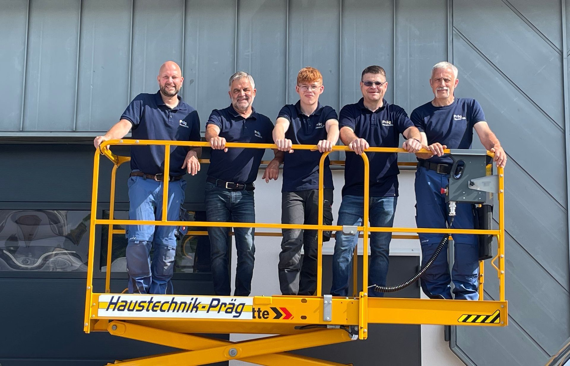 Fünf Arbeiter in passenden dunkelblauen Hemden auf einem gelben Aufzug. Sie stehen auf einer Hebebühne vor einem Gebäude
