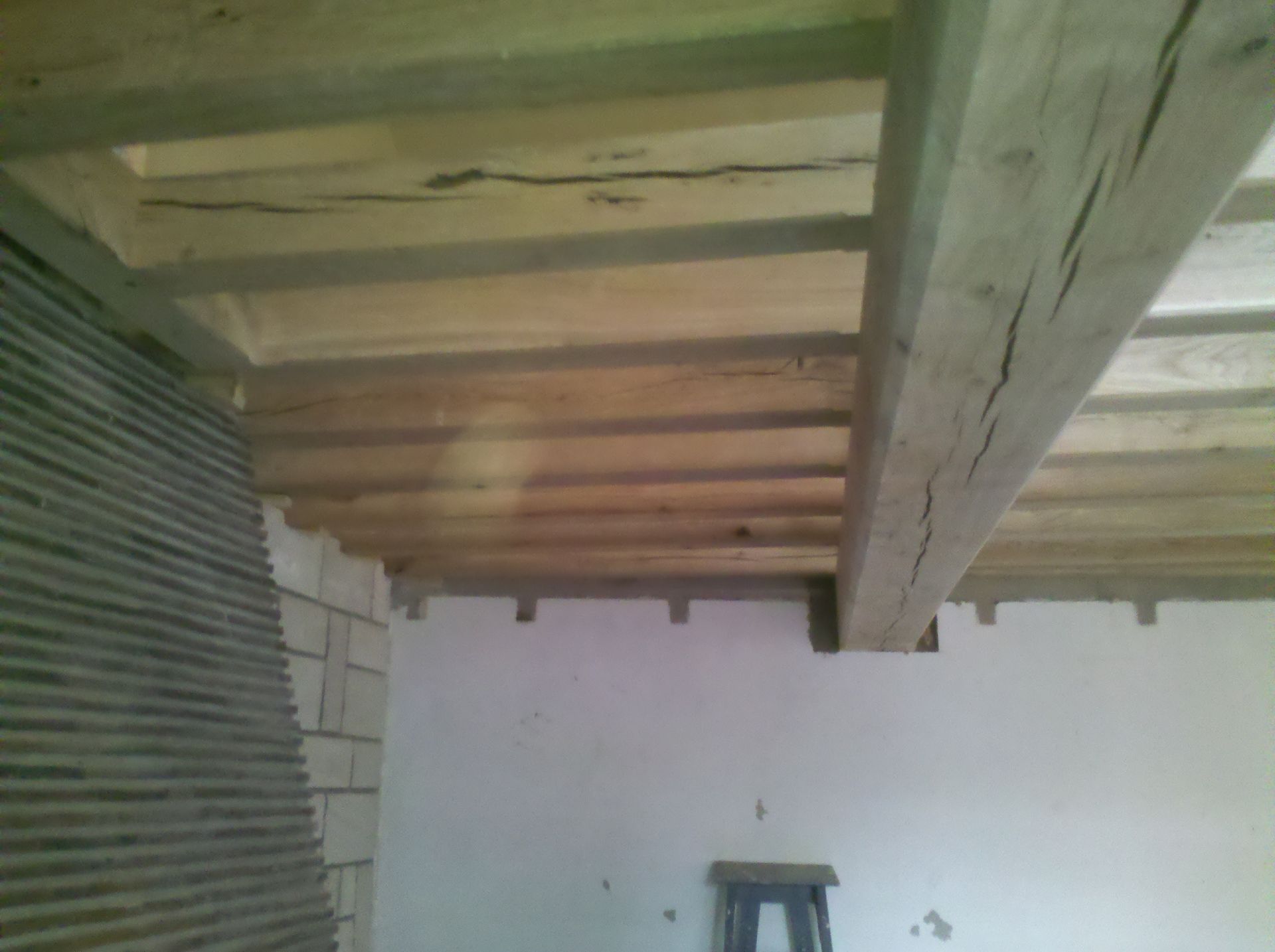 Poutres apparentes au plafond dans une pièce aux murs blancs, avec un mur en tôle ondulée à gauche