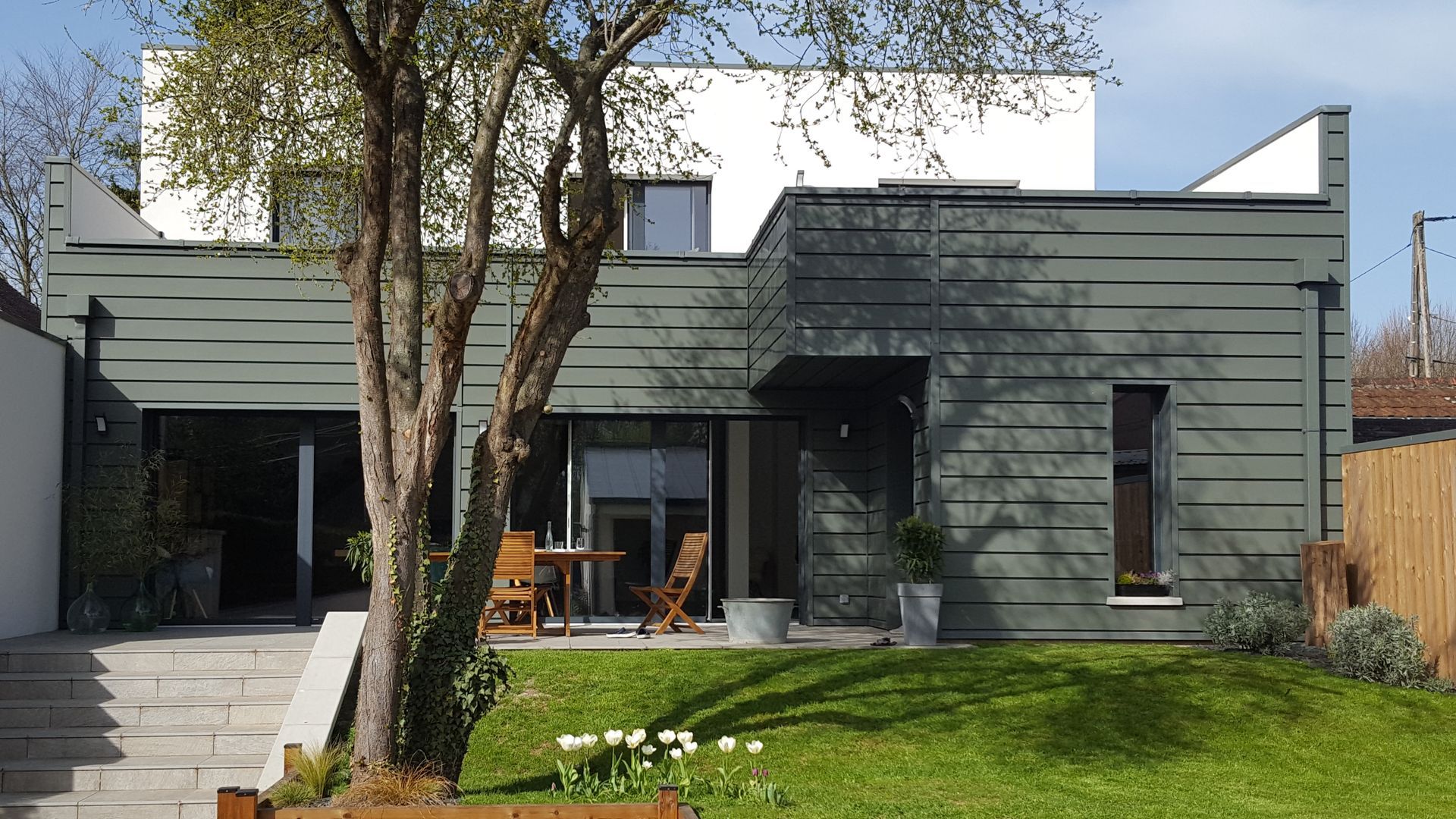 Maison moderne grise avec un petit patio, une pelouse et des arbres devant la maison