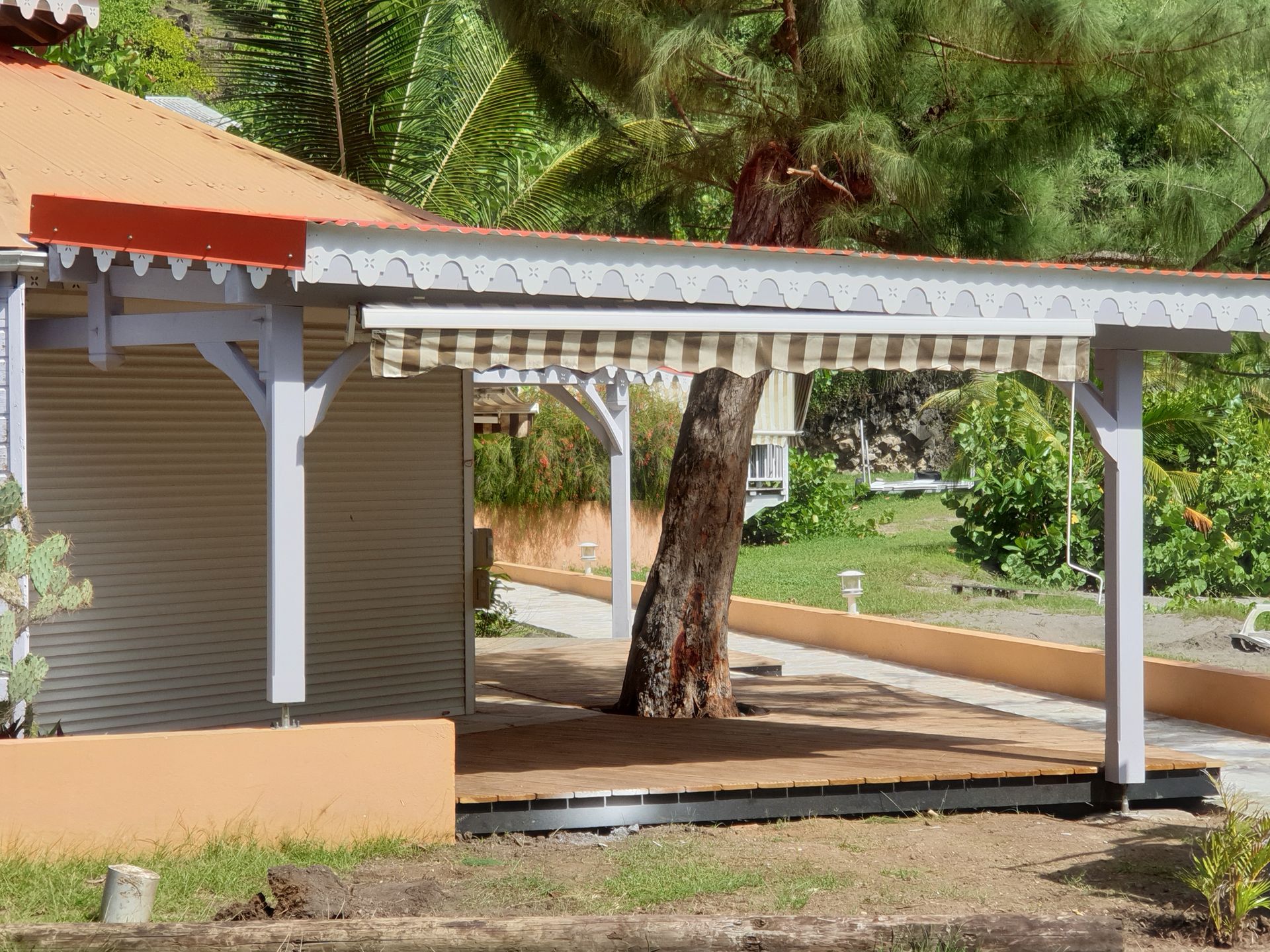 Patio couvert à côté d’un bâtiment beige, avec des colonnes blanches, un arbre et de la verdure en arrière-plan