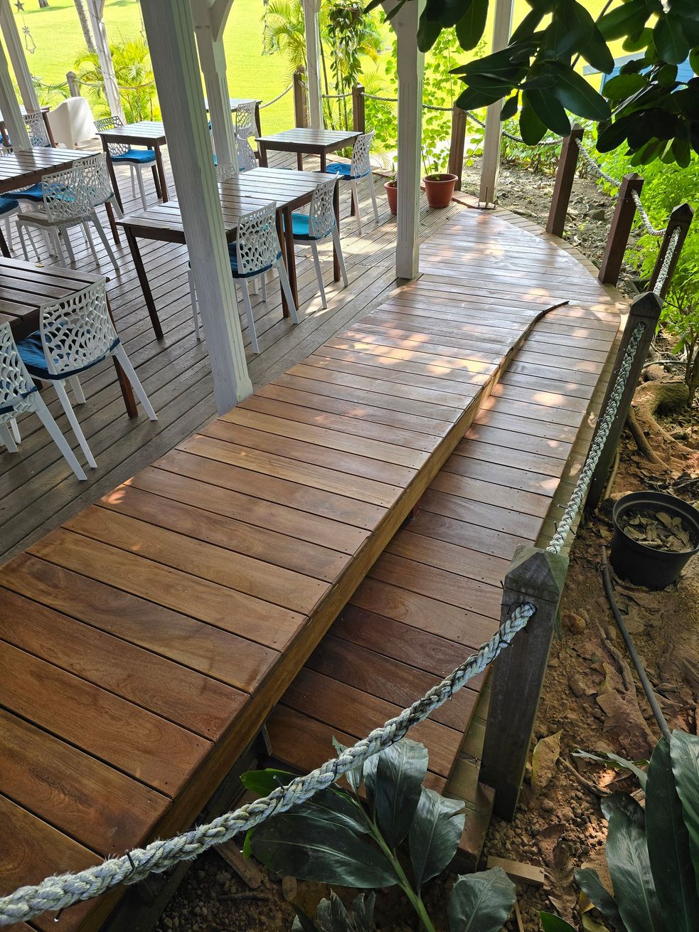 Passerelle en bois en pente à côté d’un café en plein air sous les arbres, avec des garde-corps en corde et une lumière tamisée par le feuillage.