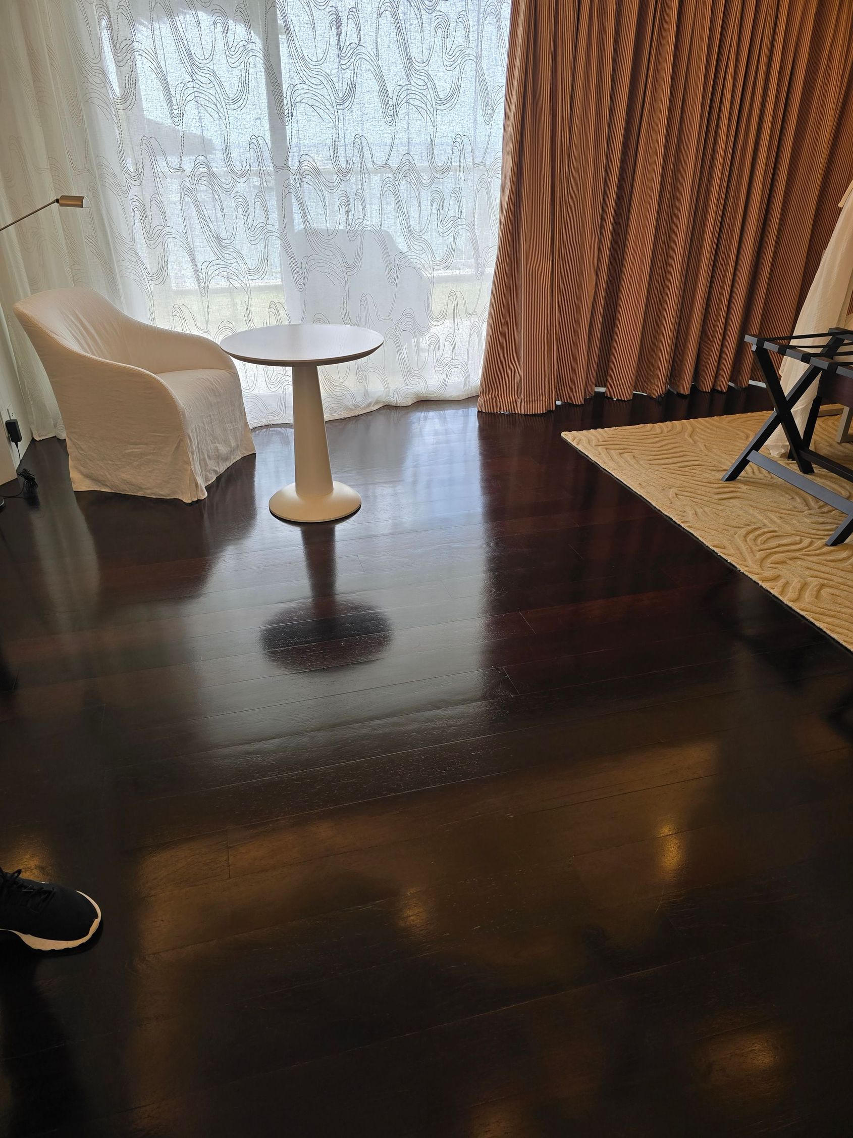Chambre d’hôtel avec une chaise blanche, une petite table ronde, des rideaux transparents et un parquet foncé.