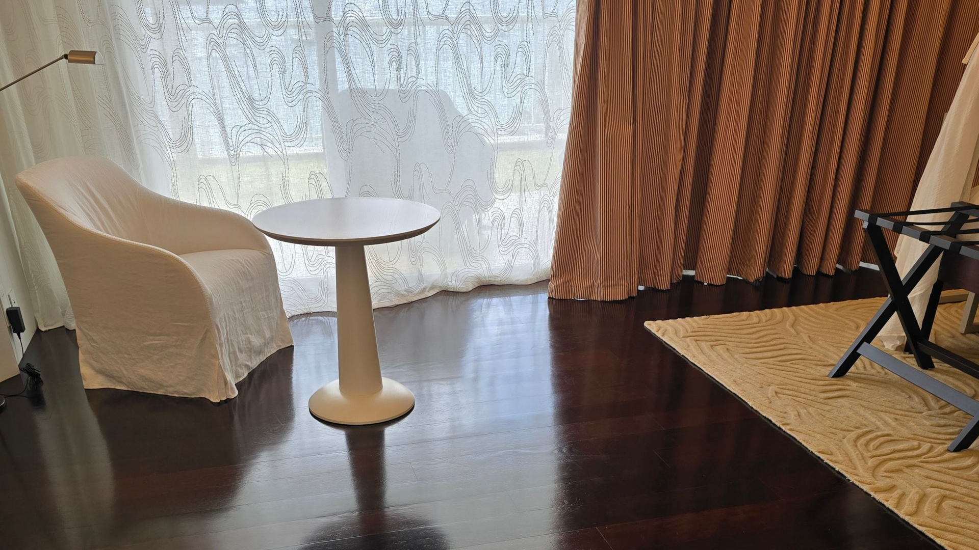 Petit salon avec un fauteuil blanc, une table d’appoint ronde, un parquet foncé et des rideaux près d’une fenêtre.