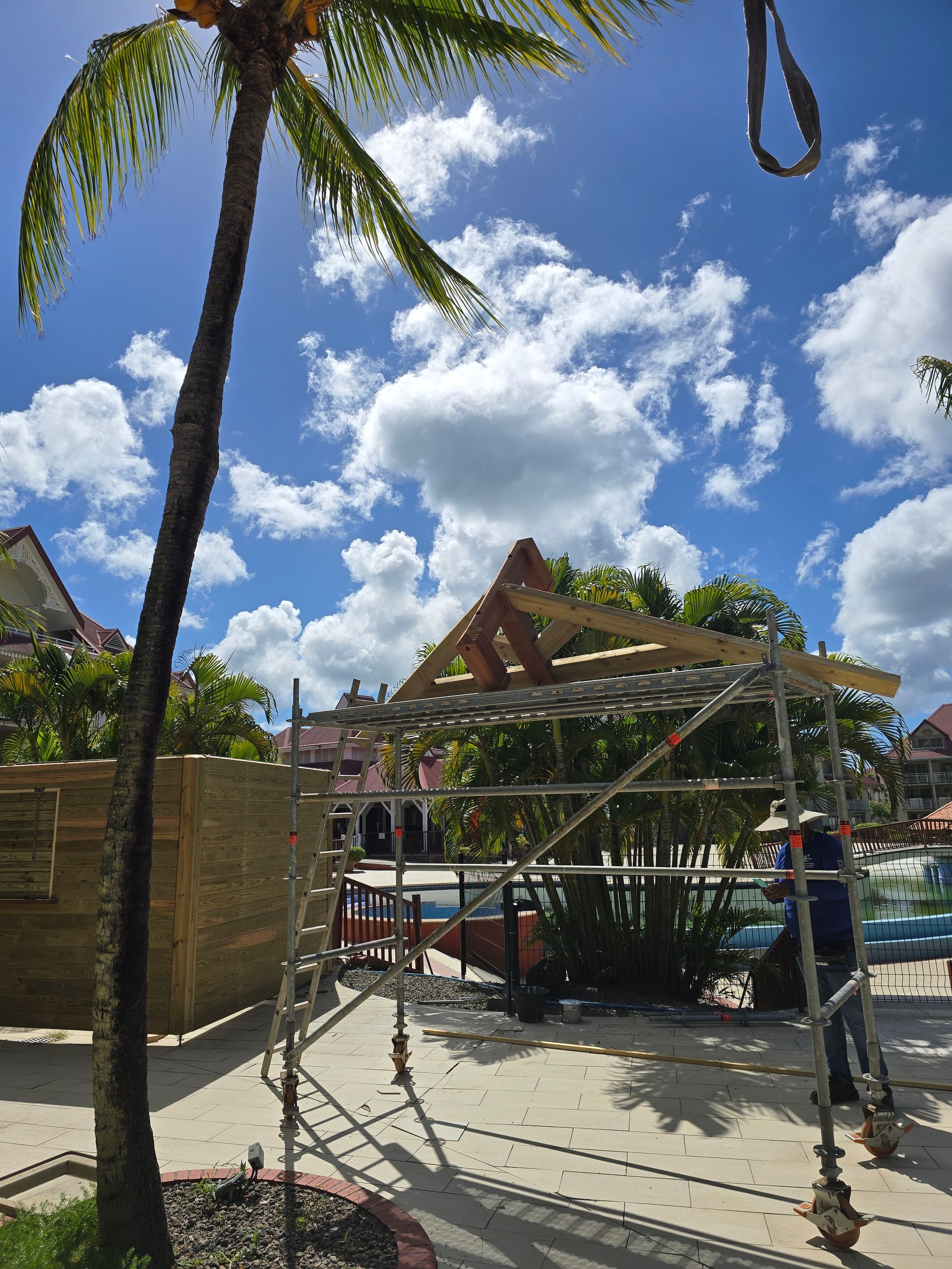 Entrée d’un complexe tropical bordée de palmiers, avec une structure au toit de chaume et un ciel bleu dégagé parsemé de nuages.