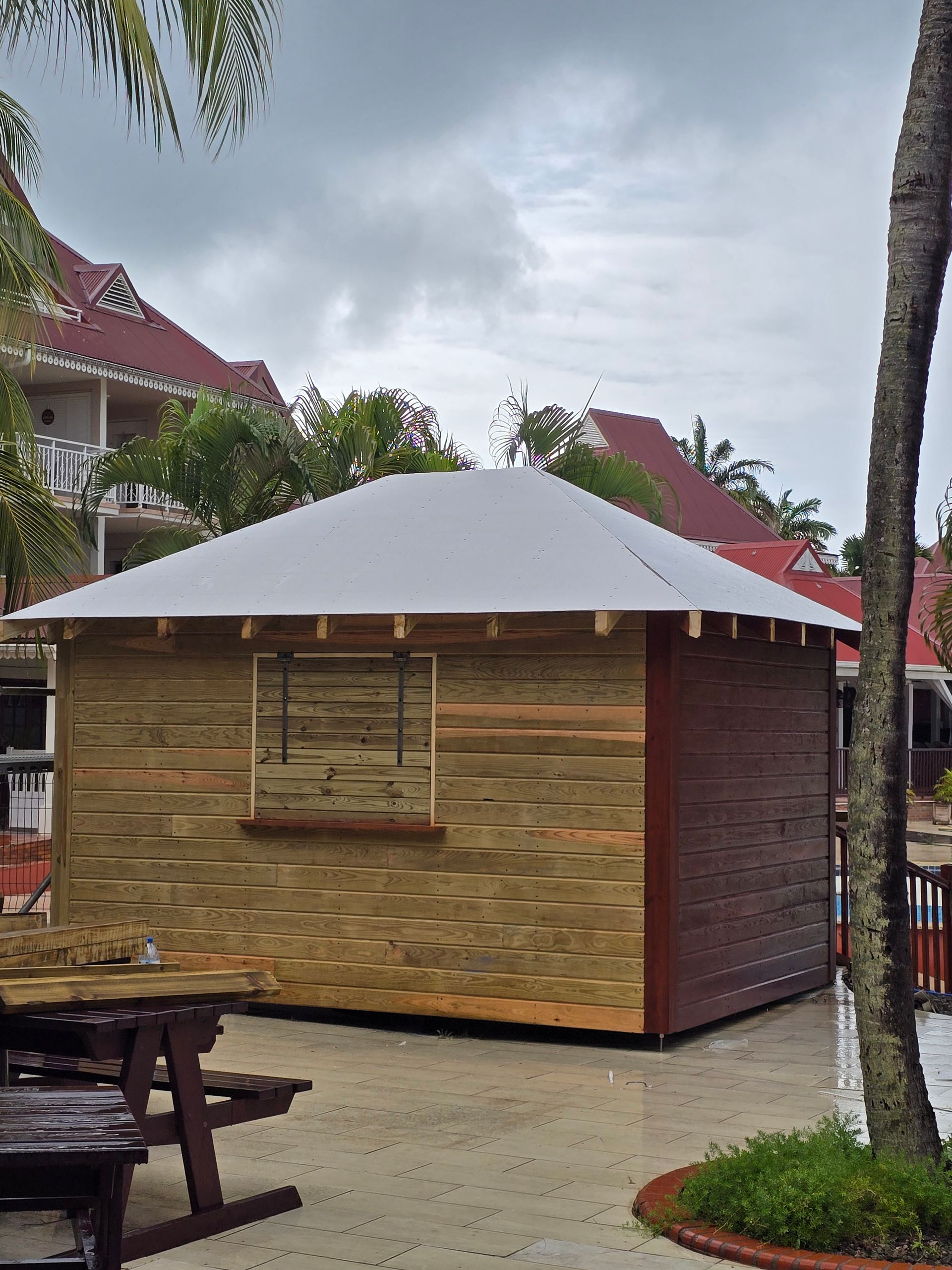 Petit pavillon en bois avec un toit blanc dans une cour tropicale, avec des palmiers et des tables de pique-nique à proximité