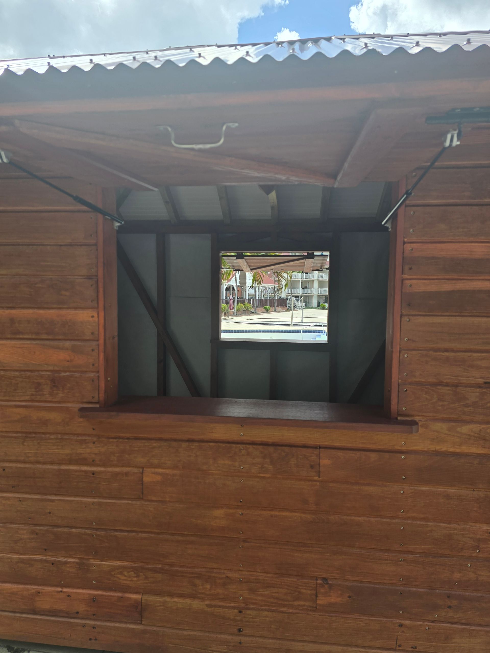 Kiosque en bois avec une fenêtre de service ouverte, sous un toit en tôle ondulée, donnant sur une rue.