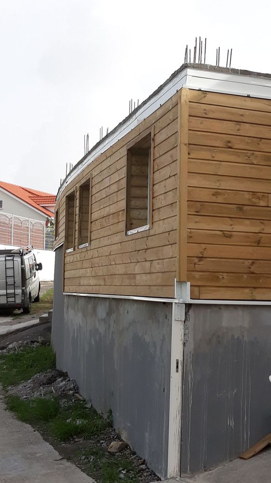 Maison en bois partiellement construite, avec des fondations en béton, à côté d’une cour étroite et d’une camionnette garée