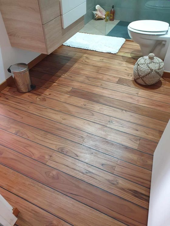 Salle de bains avec un sol en bois, des toilettes, une poubelle et un tapis de bain blanc près de la douche.