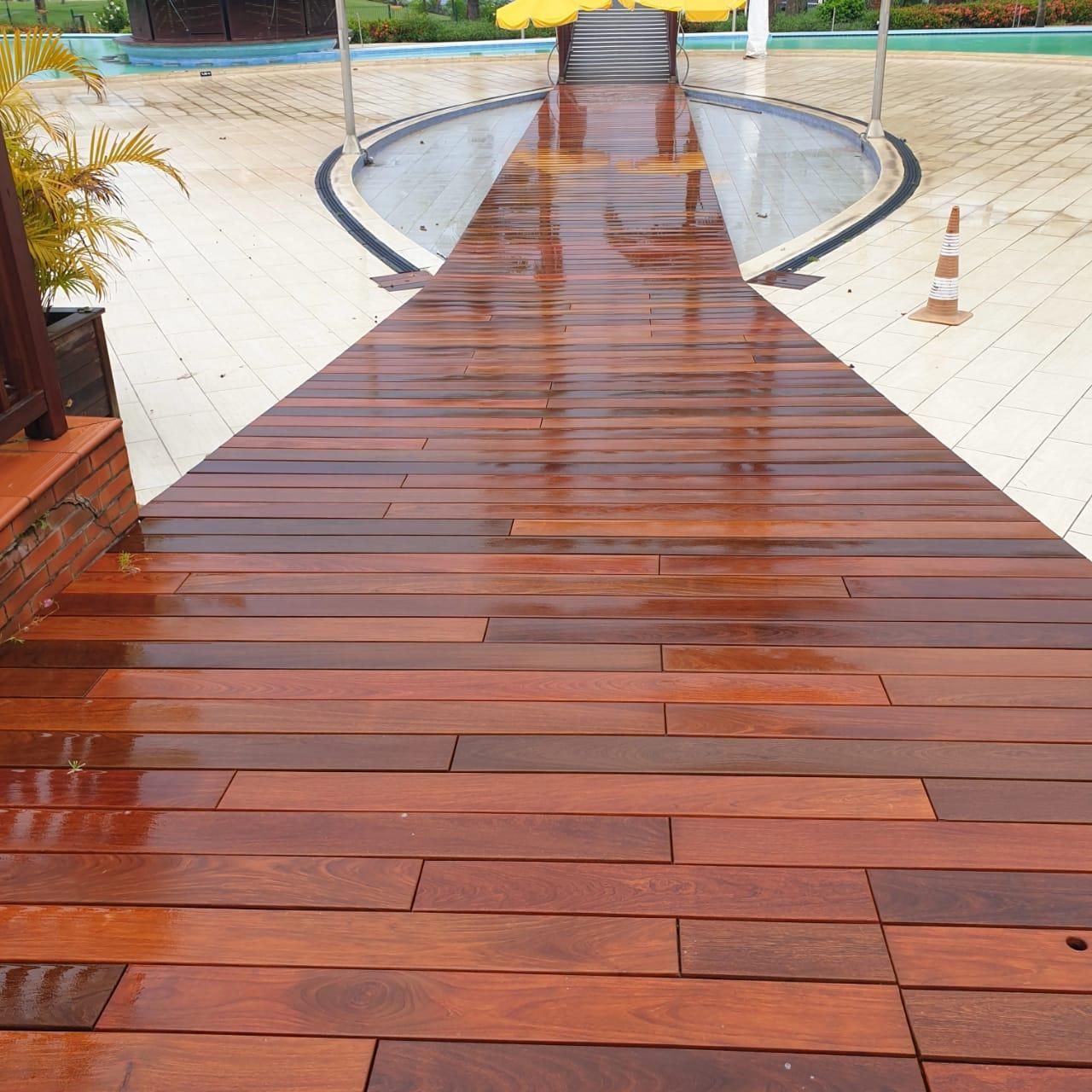 Passerelle en bois menant à une piscine, bordée de carrelage blanc et de palmiers, avec un petit cône à droite.