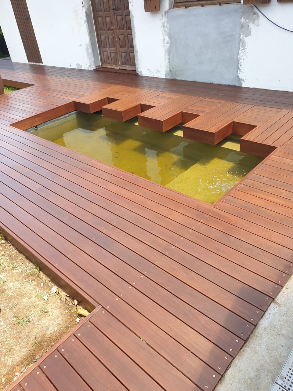 Une terrasse en bois entoure un bassin rectangulaire peu profond, agrémenté de blocs japonais et rempli d’une eau jaune-vert.