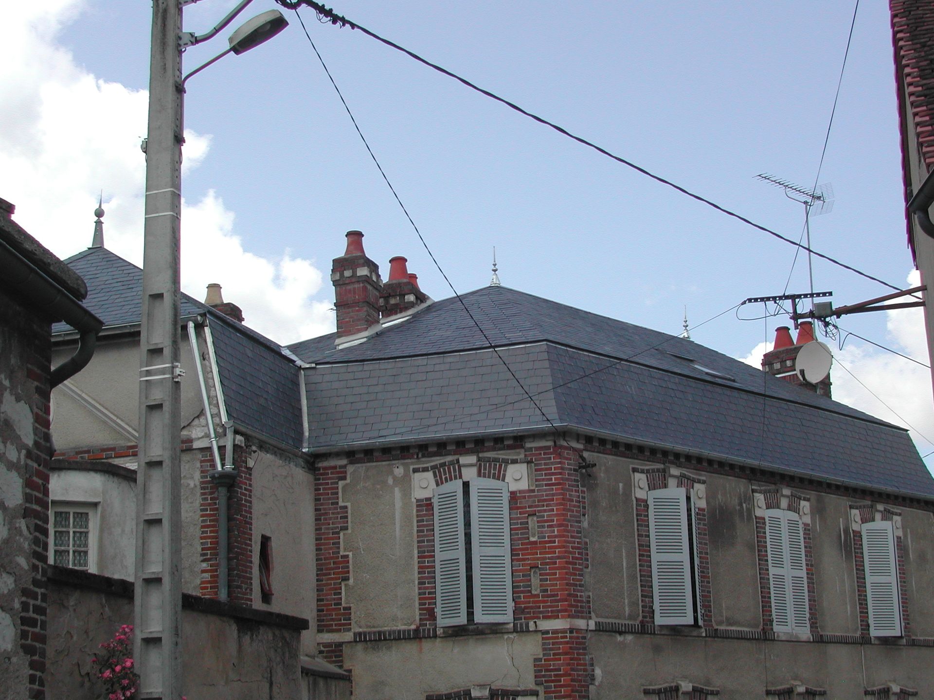 Maison en pierre avec un toit en ardoise grise, des volets blancs et des cheminées en brique, sous un ciel bleu traversé par des lignes électriques.