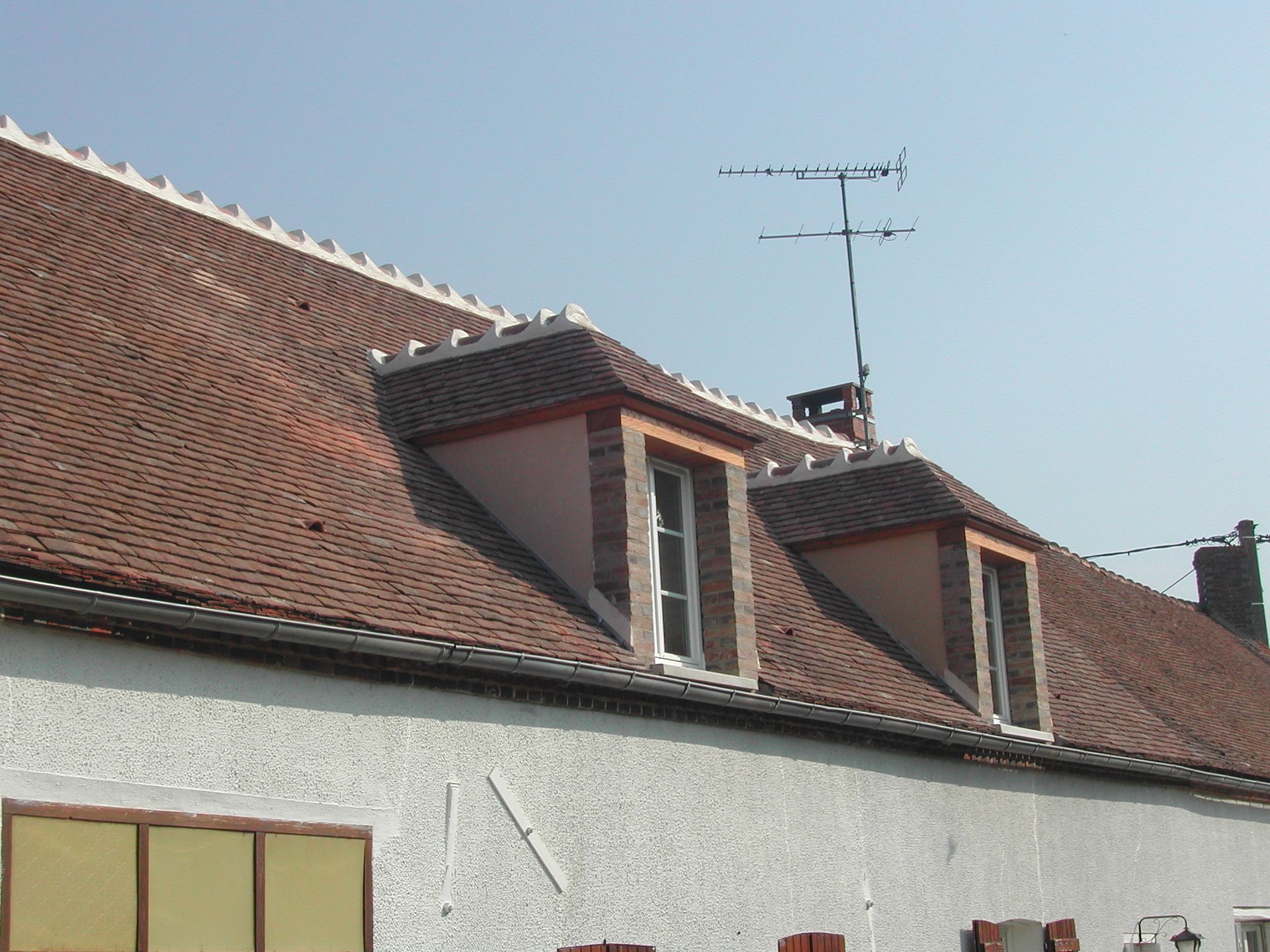 Maison blanche aux murs enduits, avec un toit de tuiles rouges et deux lucarnes, sous un ciel bleu dégagé