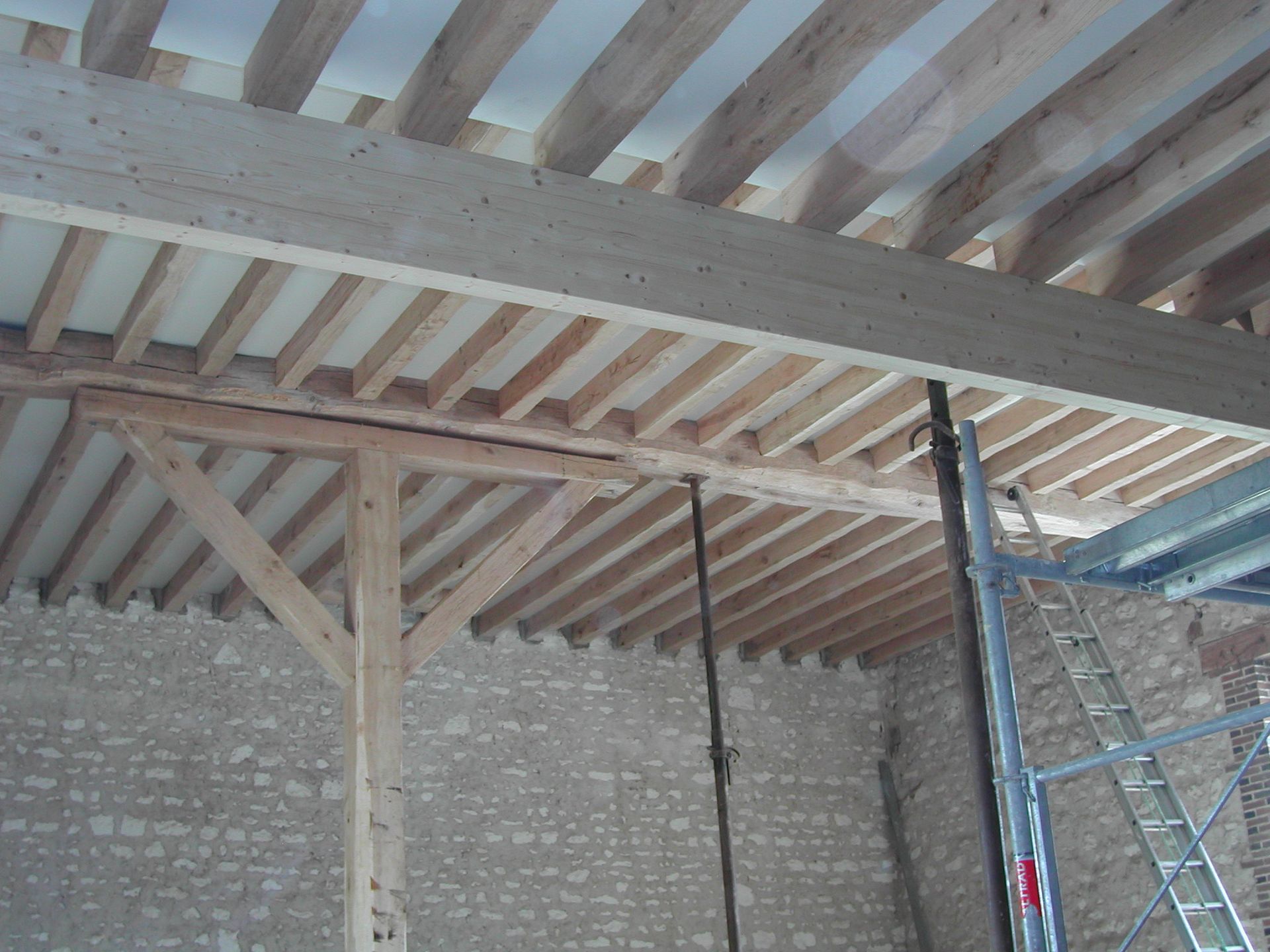 Intérieur d’un bâtiment en pierre en construction, avec des poutres en bois apparentes et une échelle appuyée contre le mur