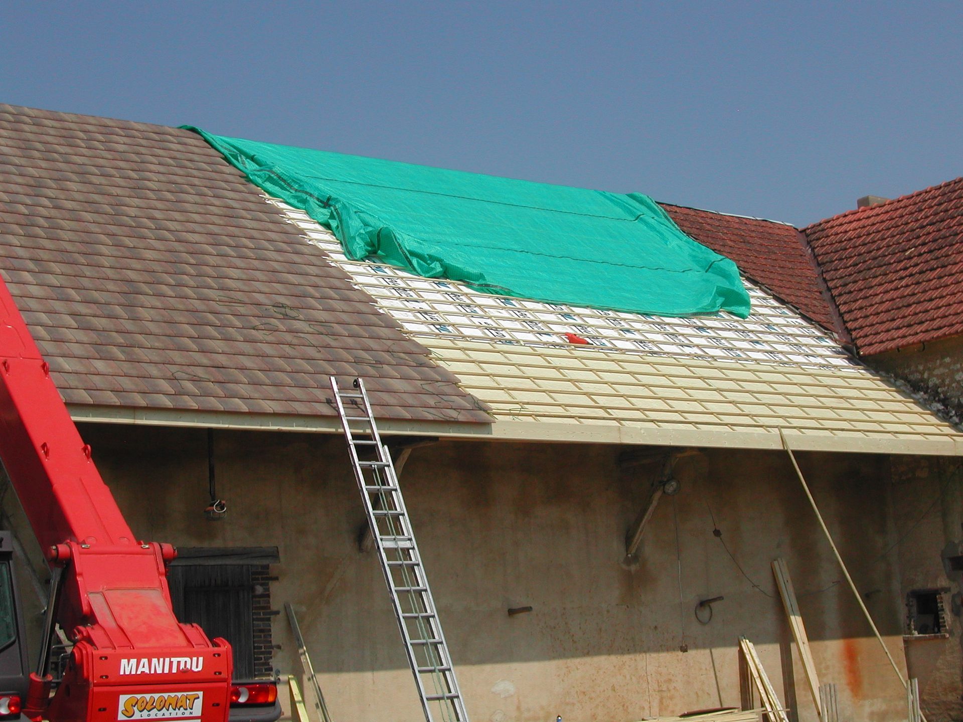 Toiture en cours de réparation, avec une bâche verte, une échelle et une grue rouge, sur une maison en construction