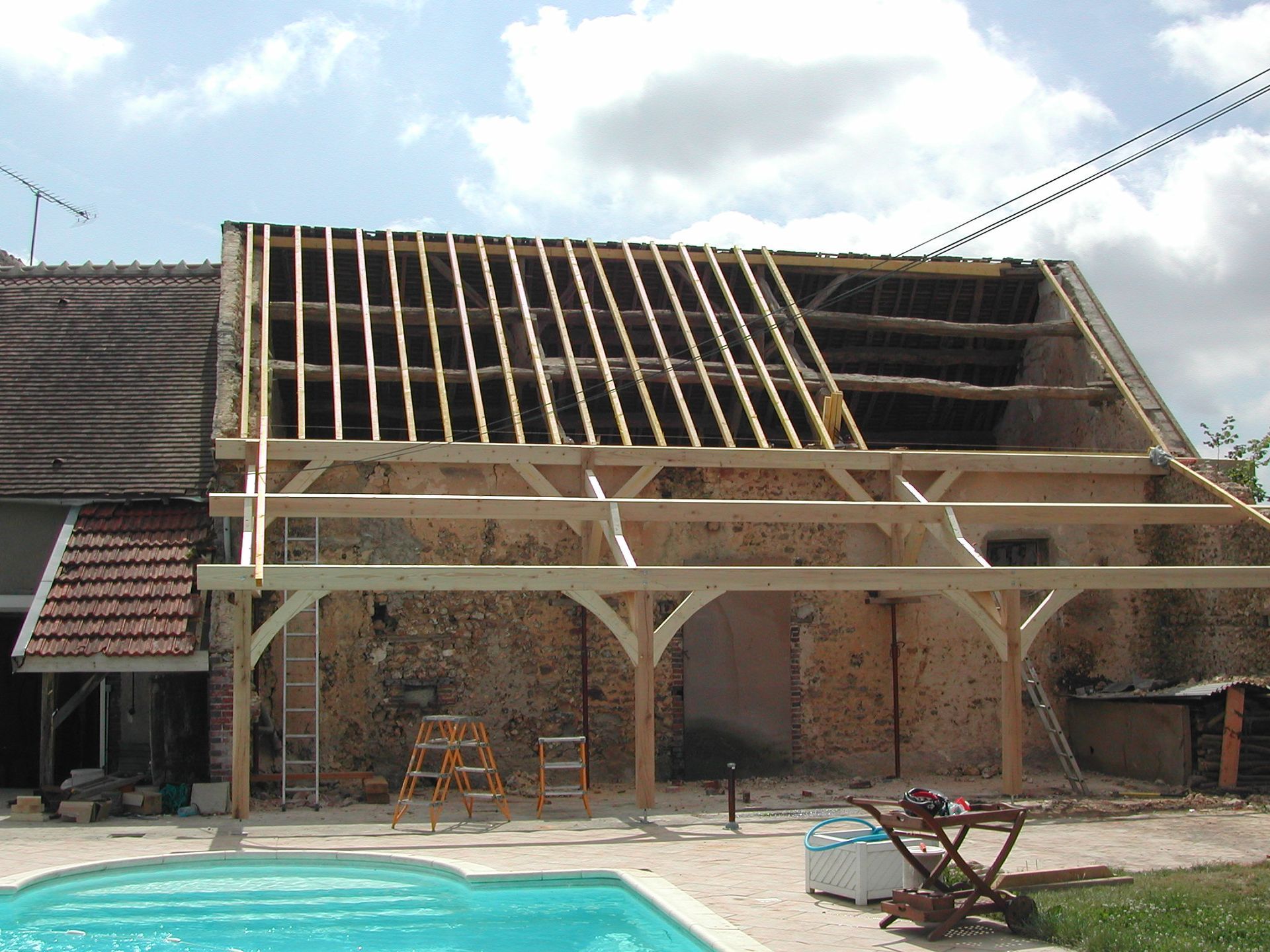 Toit d’une maison en construction, avec des chevrons en bois apparents et un échafaudage devant un bâtiment en briques.