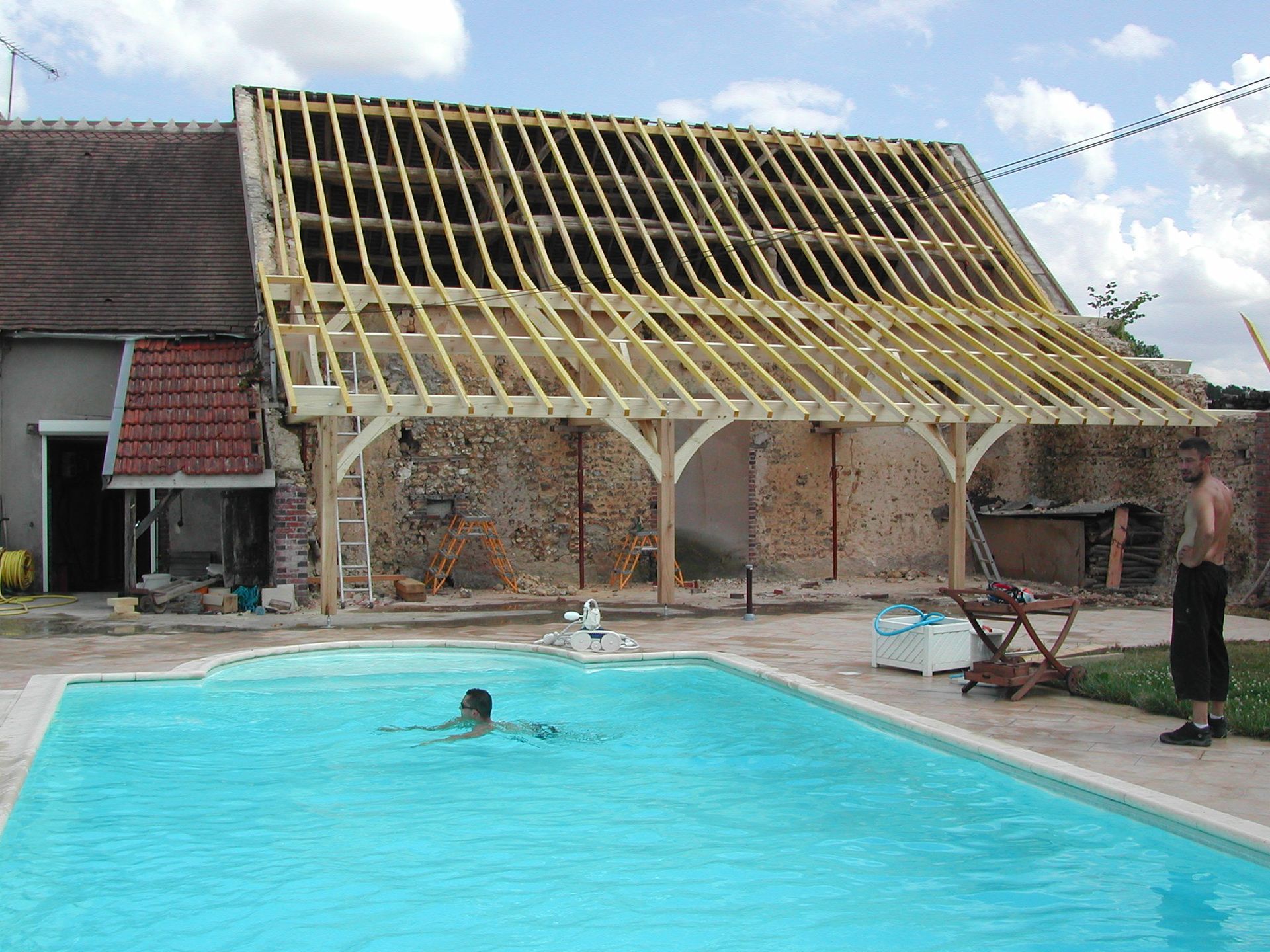 Piscine à côté d’une maison dont la toiture est en cours de rénovation, avec une personne dans l’eau et une autre au bord de la piscine.