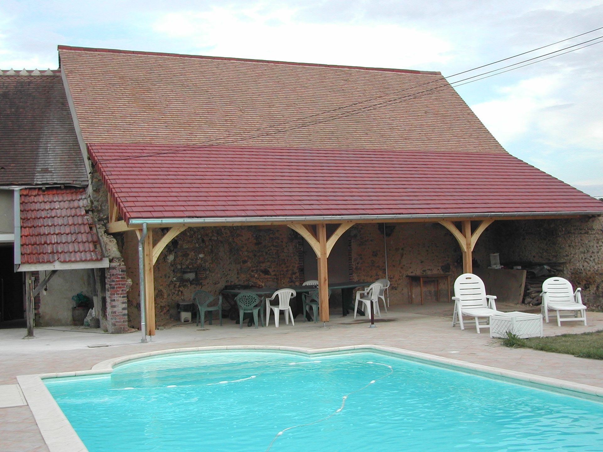 Piscine dans le jardin, à côté d’une maison rustique au toit rouge et de chaises de terrasse
