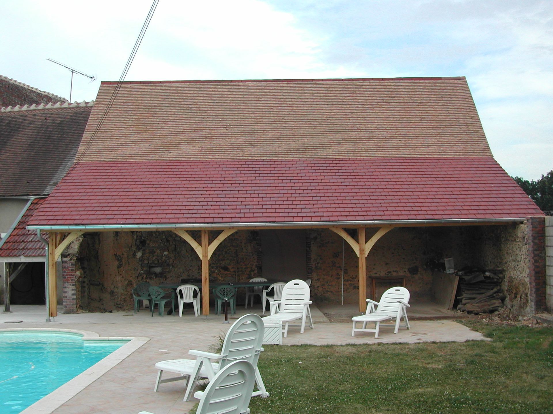 Un bâtiment rustique au toit rouge près d’une piscine, avec des chaises blanches sur la terrasse.