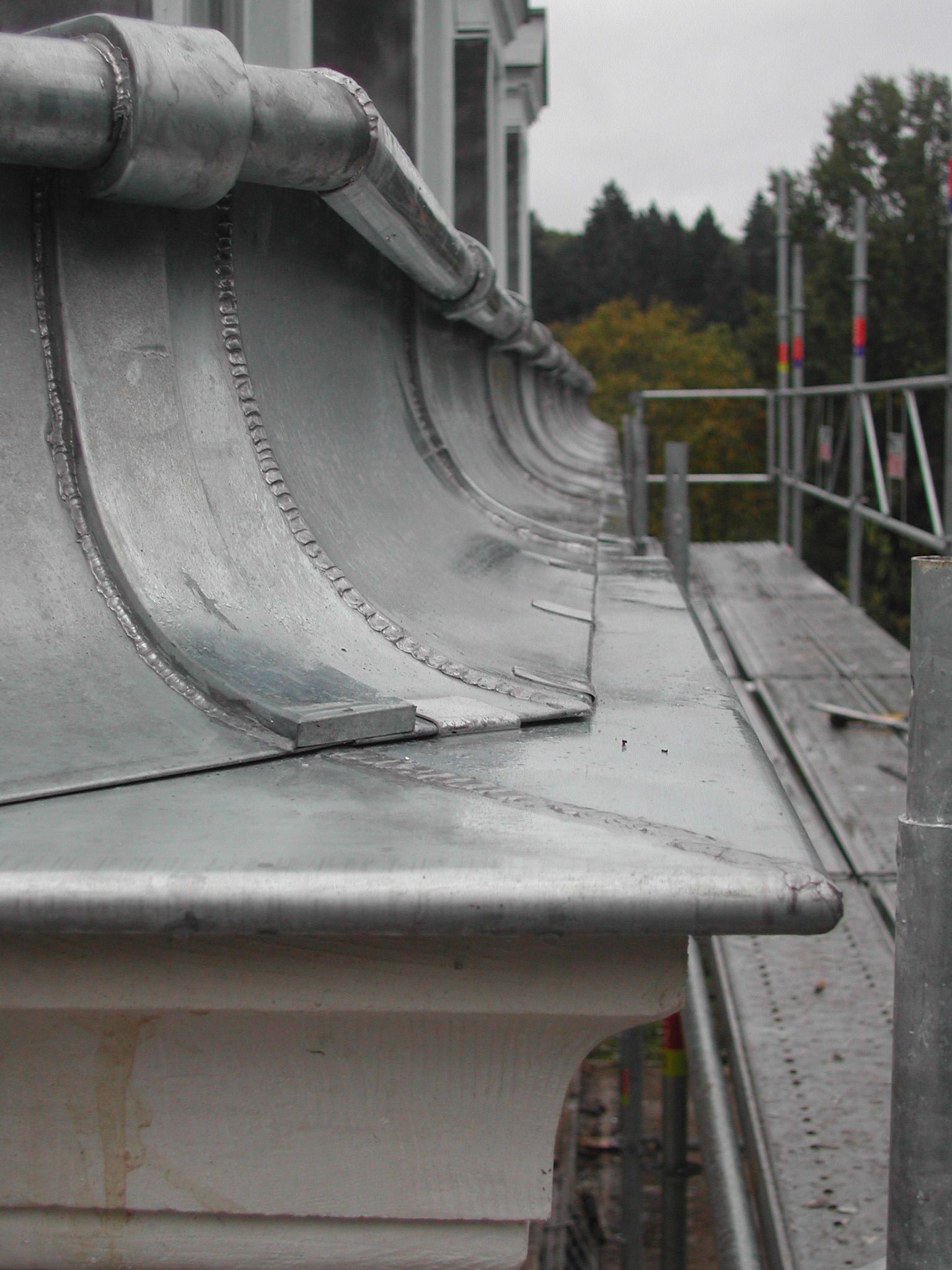 Gros plan sur le bord d’un toit en métal gris, orné de joints décoratifs et d’une rambarde, à l’extérieur sous un ciel nuageux