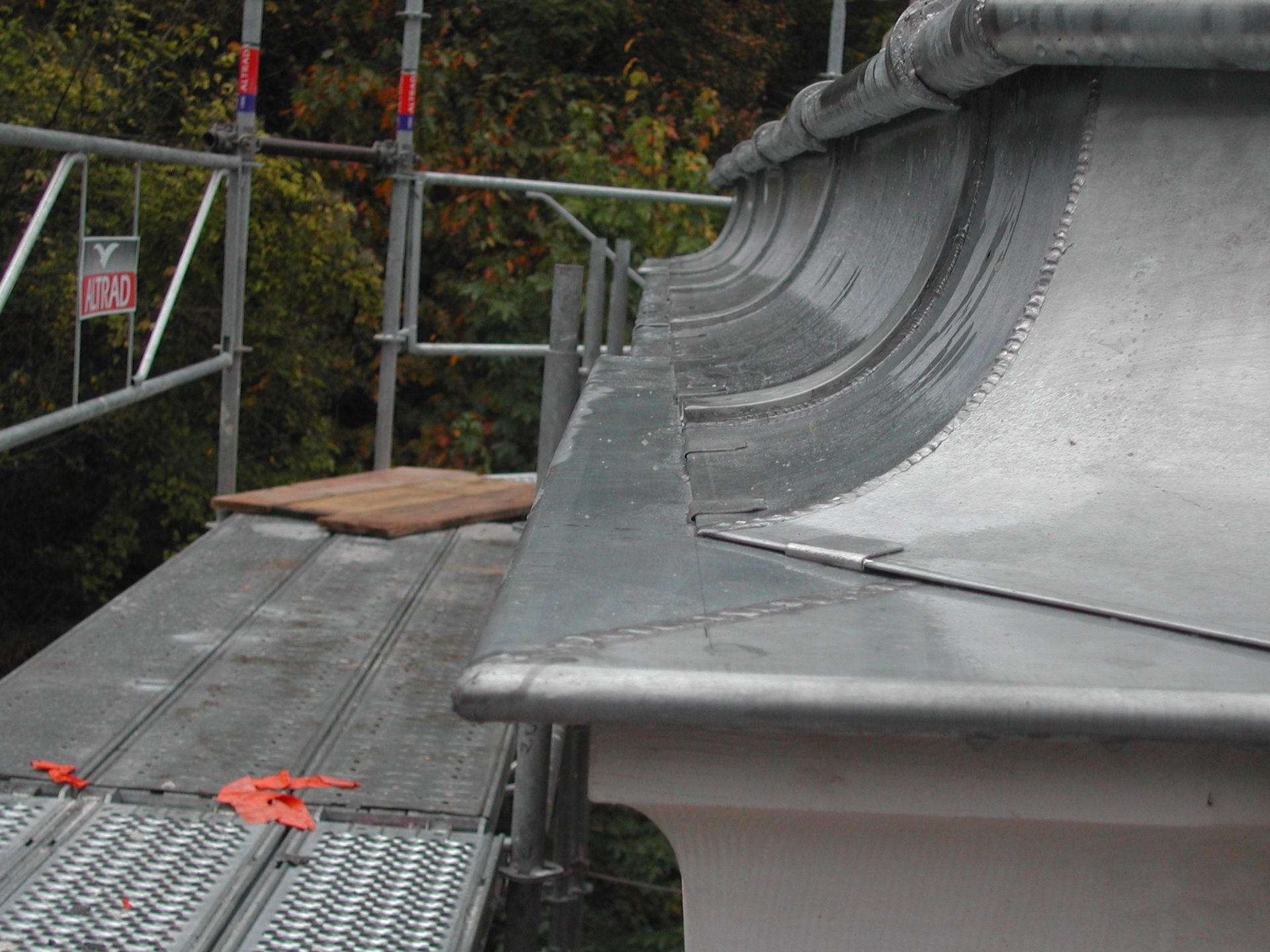 Bord de toiture métallique incurvé sur le toit d’un bâtiment entouré d’échafaudages, avec un platelage gris et des sangles de sécurité orange.