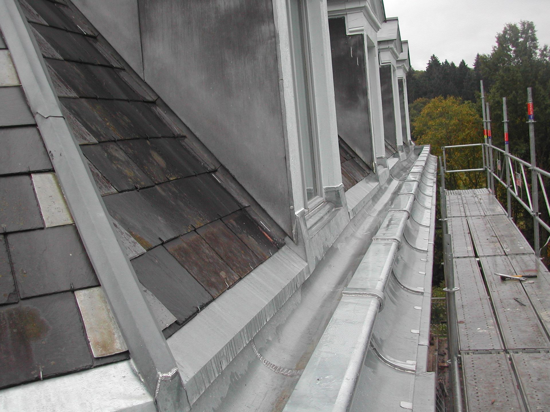 Passerelle sur le toit avec un solin métallique le long d’un bâtiment gris, à côté d’une plateforme échafaudée.