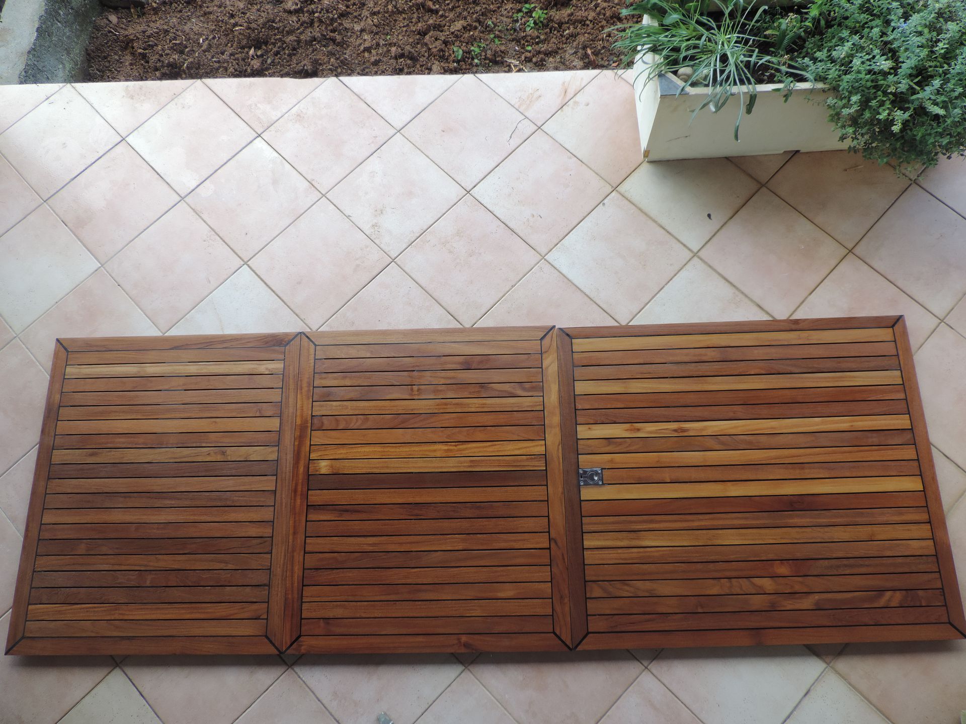 Trois panneaux à lattes en bois posés sur un carrelage beige, à côté d’une jardinière.