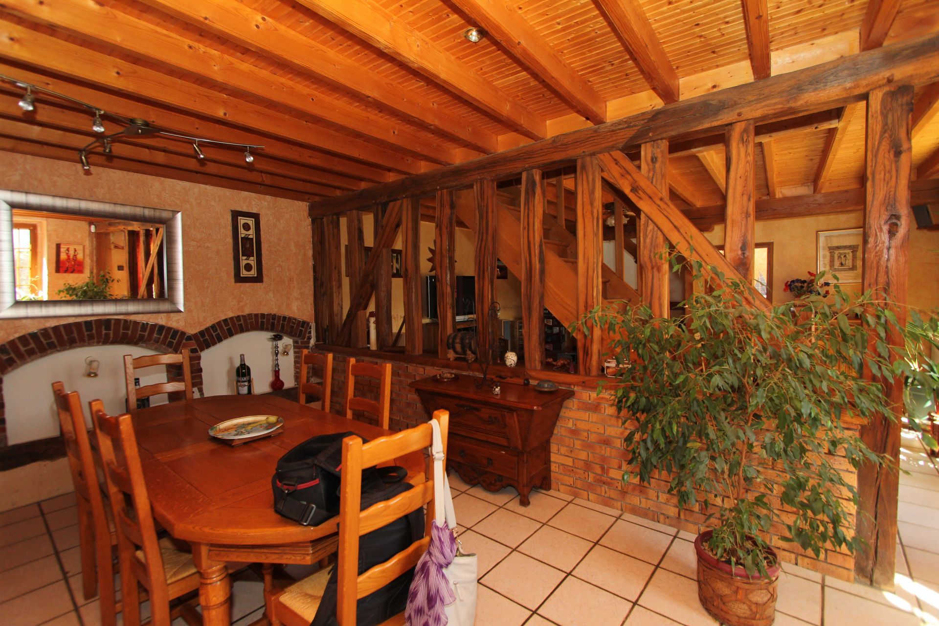 Salle à manger rustique avec poutres en bois, table ronde, chaises, sol carrelé et grande plante en pot