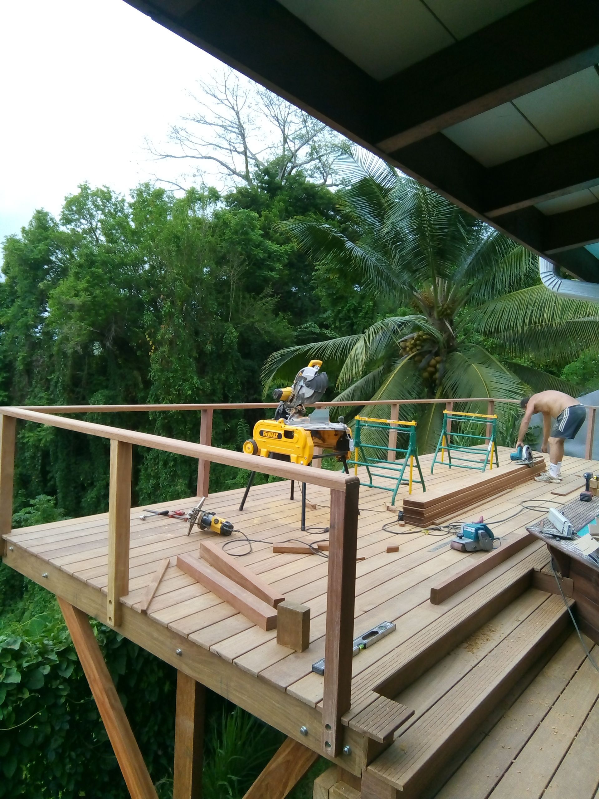 Des ouvriers construisent une terrasse en bois à flanc de colline sous les tropiques, avec des outils et du bois éparpillés autour.