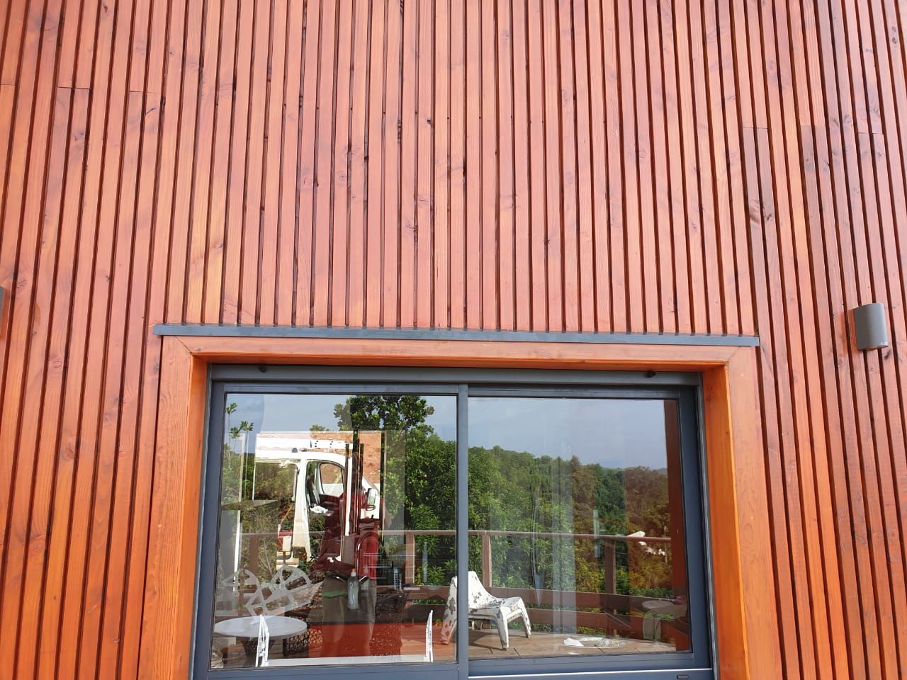 Façade de bâtiment revêtue de bois, avec une grande fenêtre reflétant des arbres et du mobilier d’extérieur