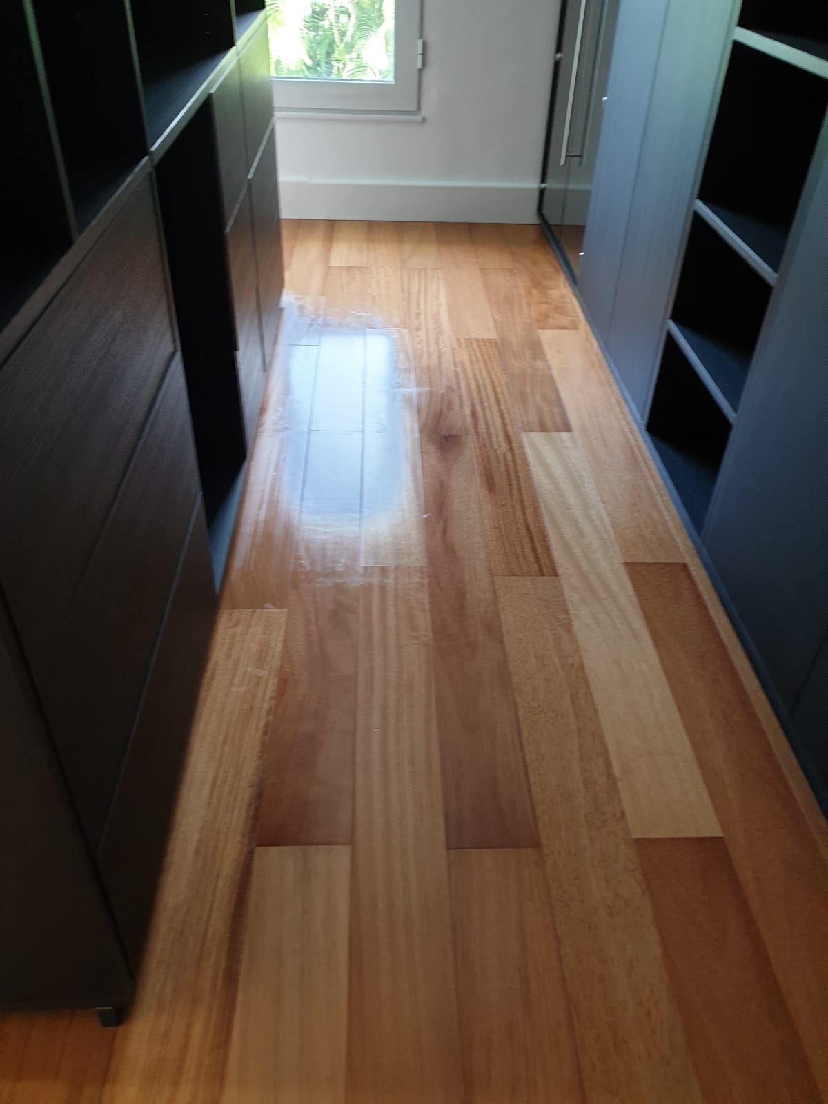 Couloir étroit avec un parquet ciré, des étagères sombres de chaque côté et une fenêtre au fond.