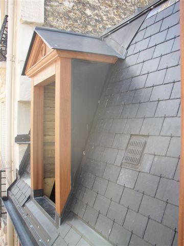 Petite lucarne avec des moulures en bois et des bardeaux d’ardoise sur un toit pentu.