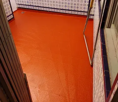 Suelo pintado de rojo en una pequeña habitación con paredes de azulejos azules y blancos estampados. Un rodillo de pintura apoyado en la pared.