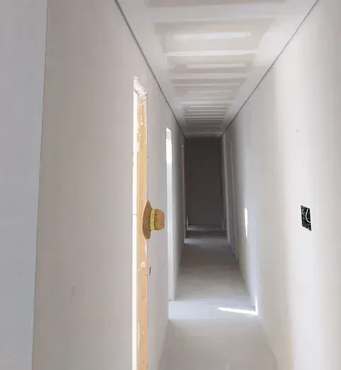 Pasillo estrecho en construcción, con paredes y techo blancos. Un sombrero cuelga de una puerta abierta.