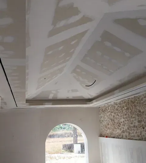Vista interior de una habitación con un techo de yeso parcialmente terminado. Incluye una puerta arqueada, un muro de piedra vista y una ventana que da al exterior.