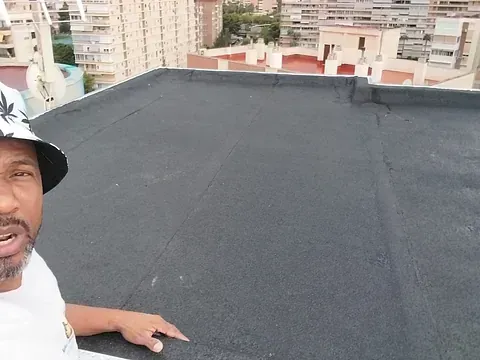 Un hombre en una azotea plana y negra con edificios urbanos al fondo. Lleva camisa blanca y sombrero.
