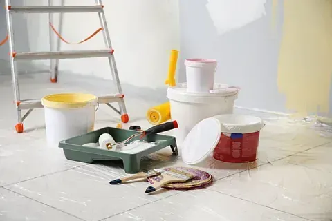 Suministros de pintura y una escalera en una habitación con paredes parcialmente pintadas, lista para renovar.