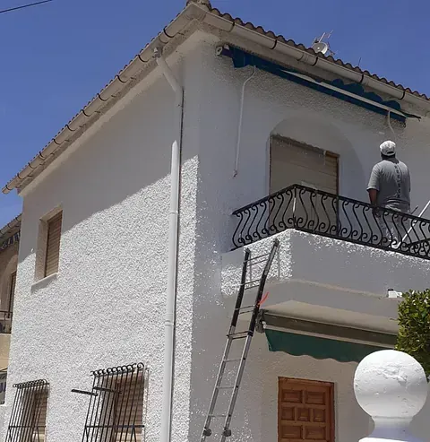 Casa blanca de dos pisos con una persona en un balcón, de pie sobre una escalera. Cielo azul al fondo.