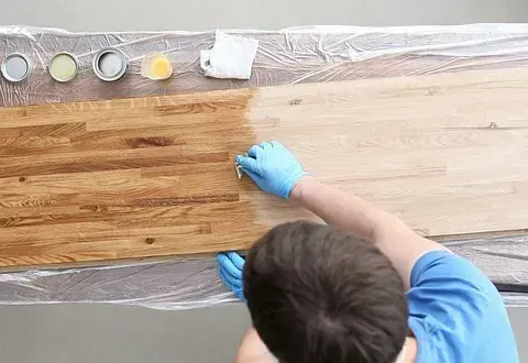 Persona aplicando tinte a una superficie de madera, con la parte teñida de un marrón más oscuro. Cerca hay varios colores de tinte y herramientas de aplicación.