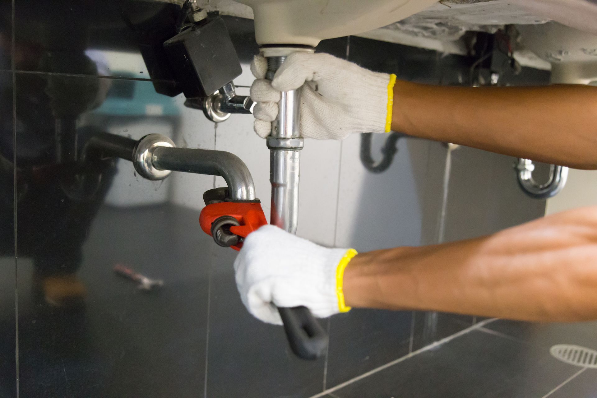 Des mains gantées utilisent une clé pour réparer un tuyau d'évier sous un comptoir.