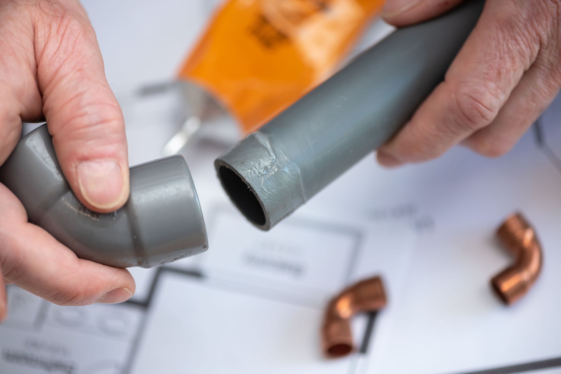 Des mains raccordent des tuyaux en PVC gris lors d'une réparation de plomberie, avec des coudes en cuivre à proximité.