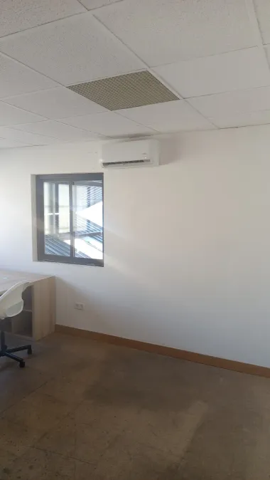Sala de oficina vacía con paredes blancas, una ventana, aire acondicionado montado en la pared, alfombra marrón y una esquina de escritorio.