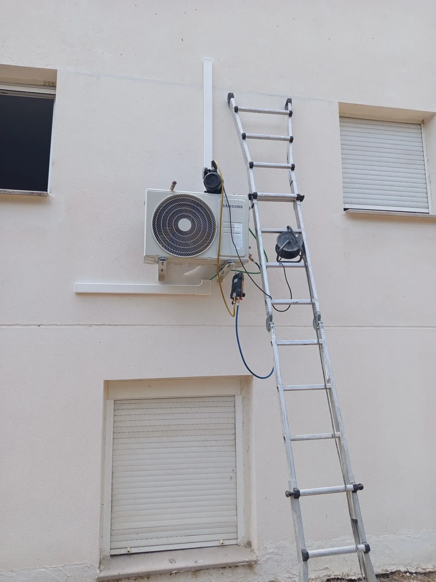 Escalera extensible apoyada en un edificio, junto a una unidad de aire acondicionado exterior y unas contraventanas.