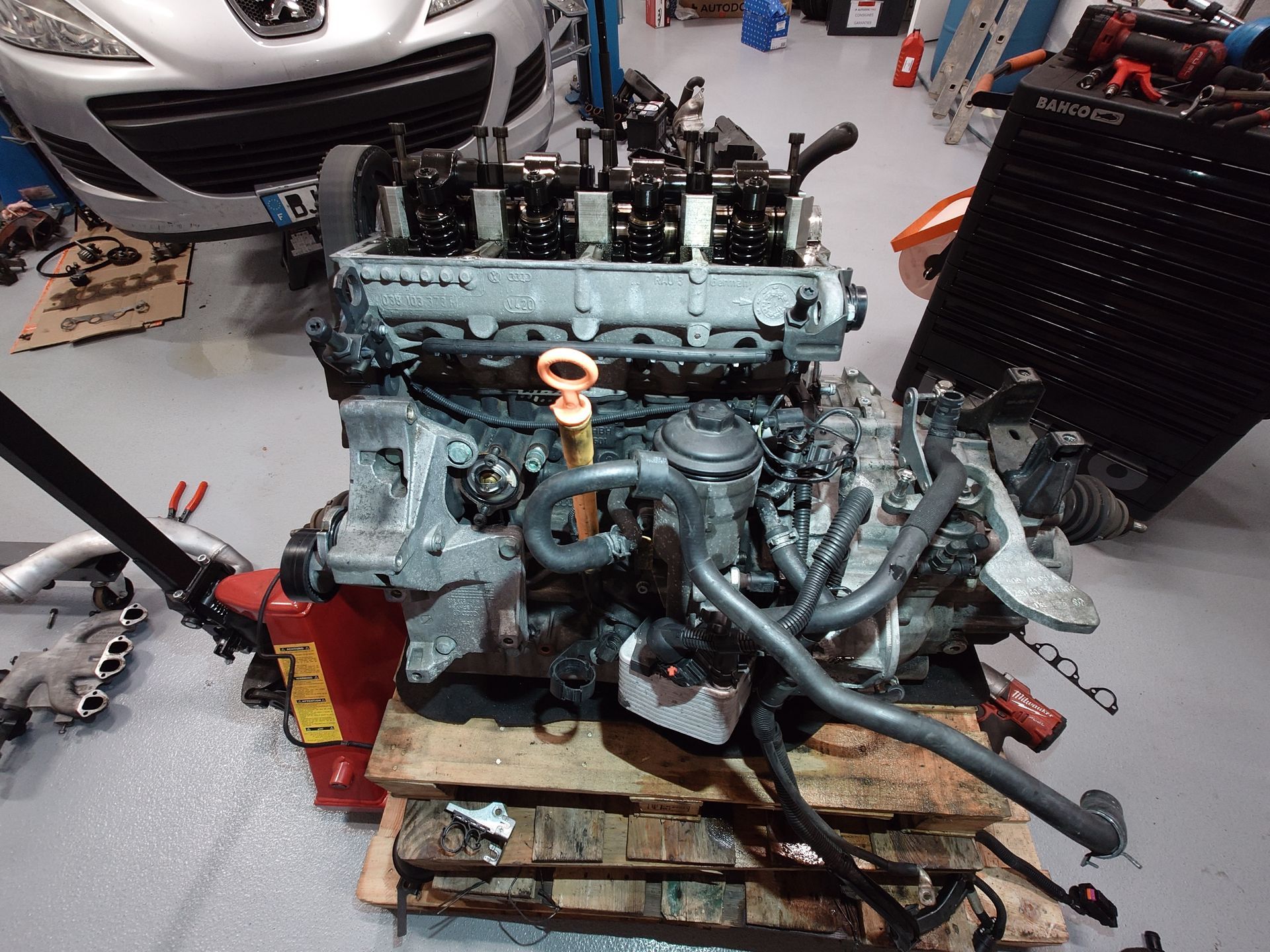 Moteur de voiture sur une palette en bois dans un garage, avec des outils et une voiture en arrière-plan.