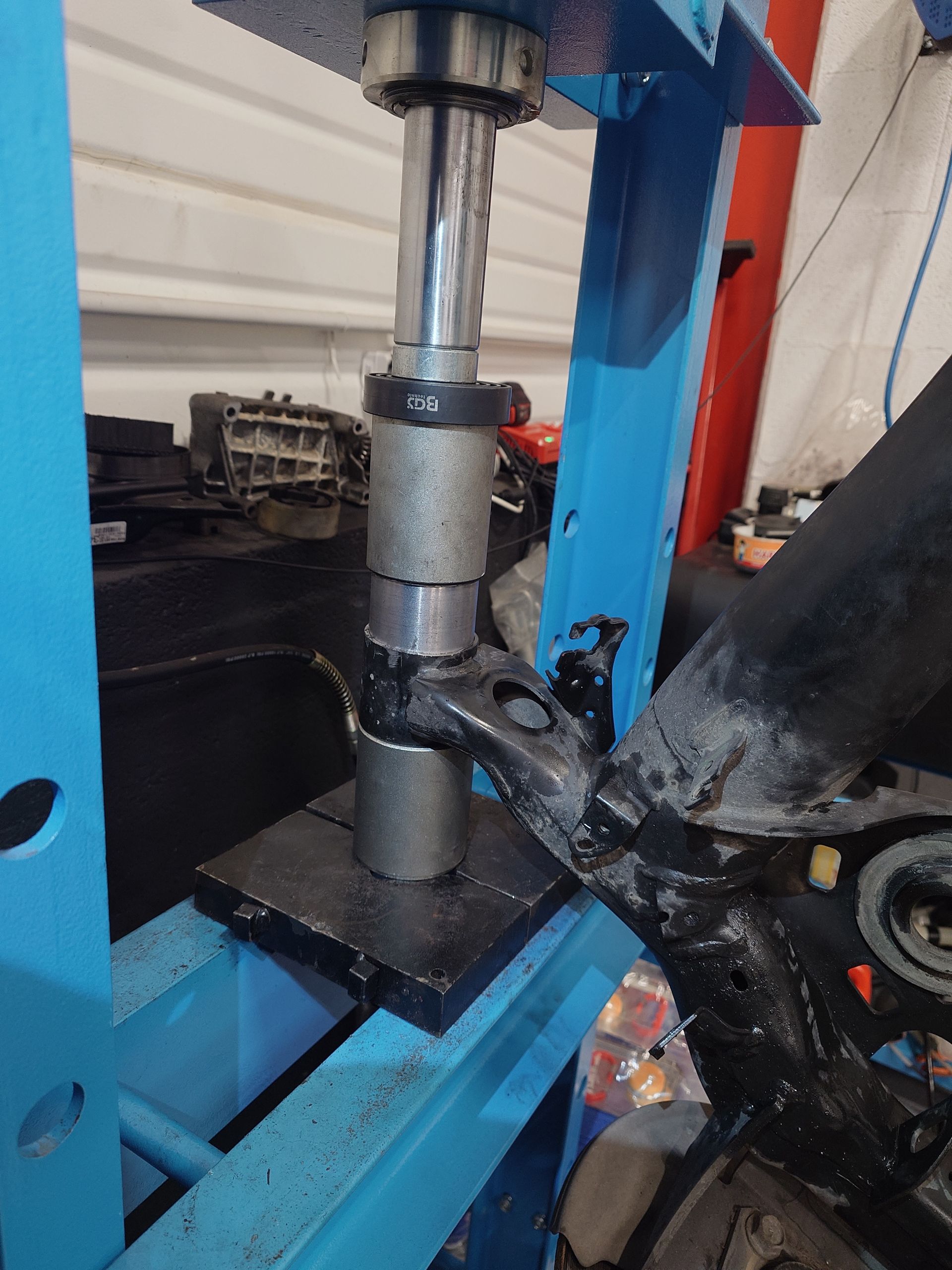 Une pièce de suspension de véhicule noire pressée dans une presse hydraulique bleue dans un garage.