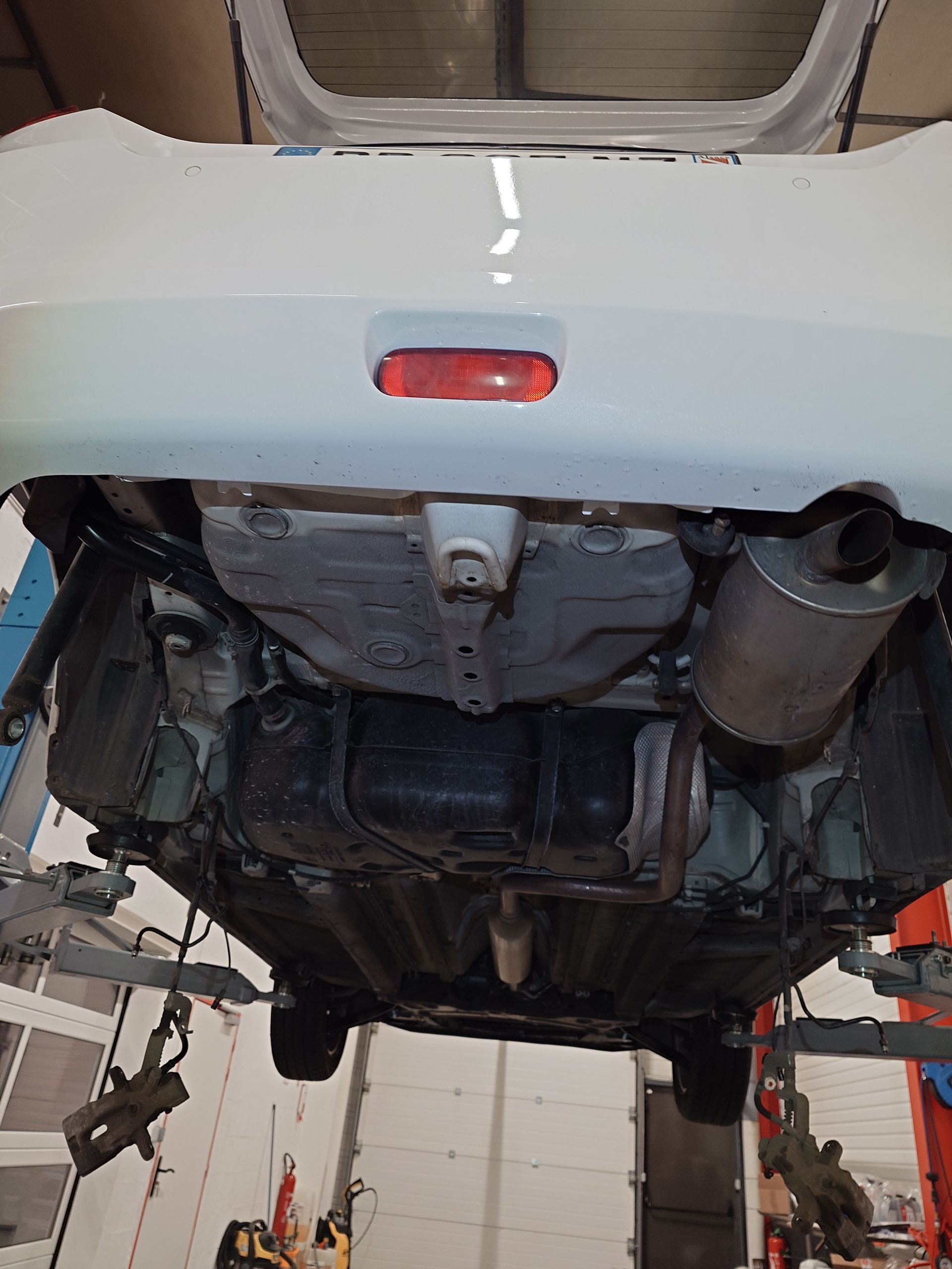 Dessous d'une voiture blanche sur un pont élévateur. On distingue les éléments noirs et le réservoir de carburant.