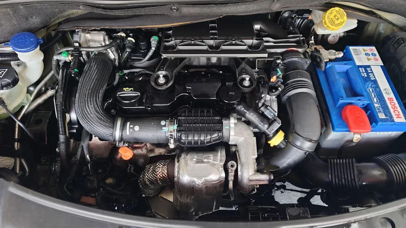 Compartiment moteur de voiture avec des durites noires, une batterie bleue à droite et un bouchon de liquide jaune en haut à droite