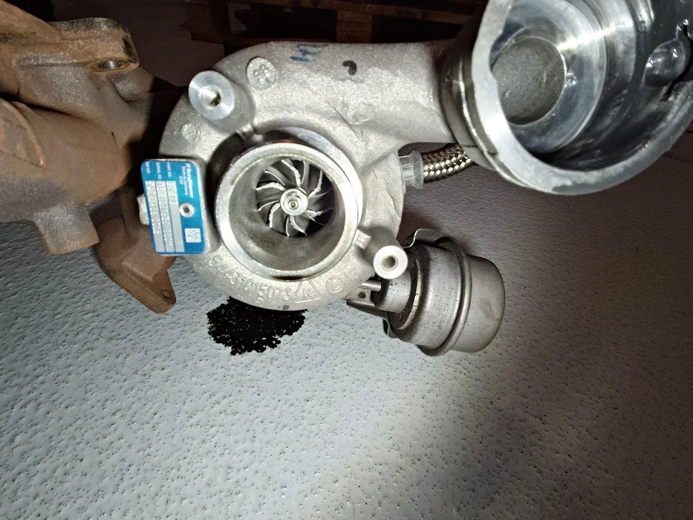 Gros plan sur un ensemble turbocompresseur de voiture posé sur un sol gris, avec des tuyaux métalliques et une étiquette bleue